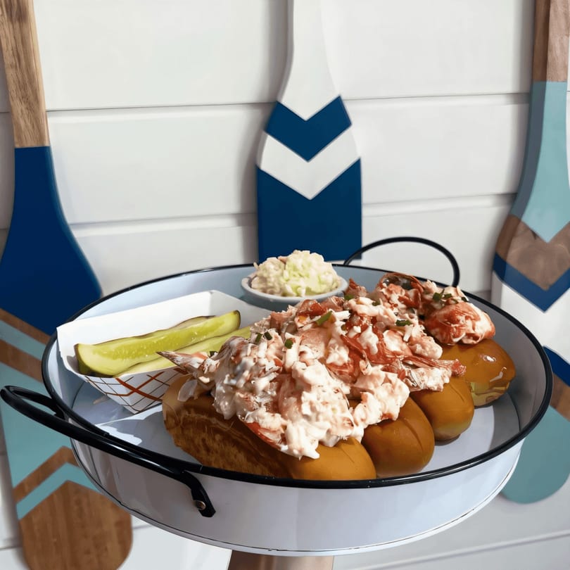 Two Mystic Lobster rolls with coleslaw and pickles on a white tray, against a nautical-themed wall.