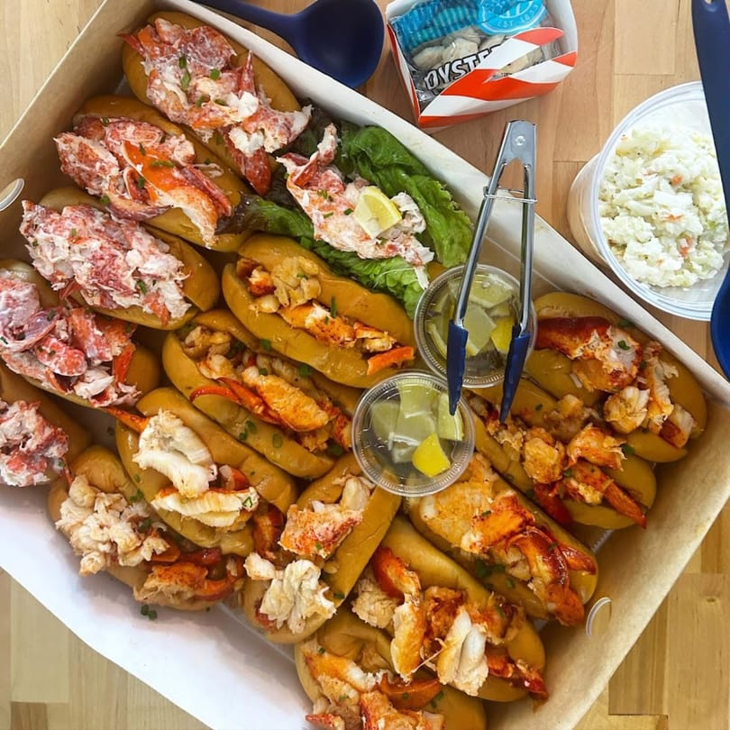 A catering box filled with Mystic Lobster Roll Co.'s lobster rolls, coleslaw, and oysters.