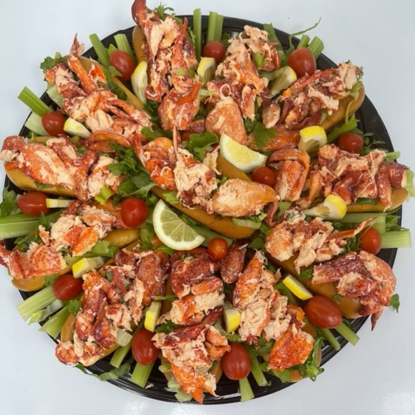 A platter of Mystic Lobster Roll Co. lobster rolls garnished with lettuce, celery, cherry tomatoes, and lemon wedges.