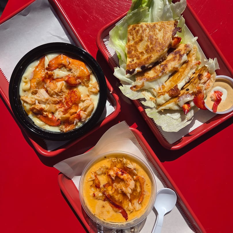 Three Mystic Lobster Co. dishes on red trays: lobster mac and cheese, lobster quesadilla, and lobster bisque.